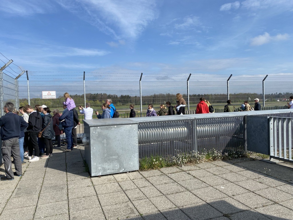 Aufnahme der Besucherterasse vom Bodensee-Airport Friedrichshafen. Zu sehen sind einige Menschen die vor einem Zaun stehen und auf das Vorfeld schauen