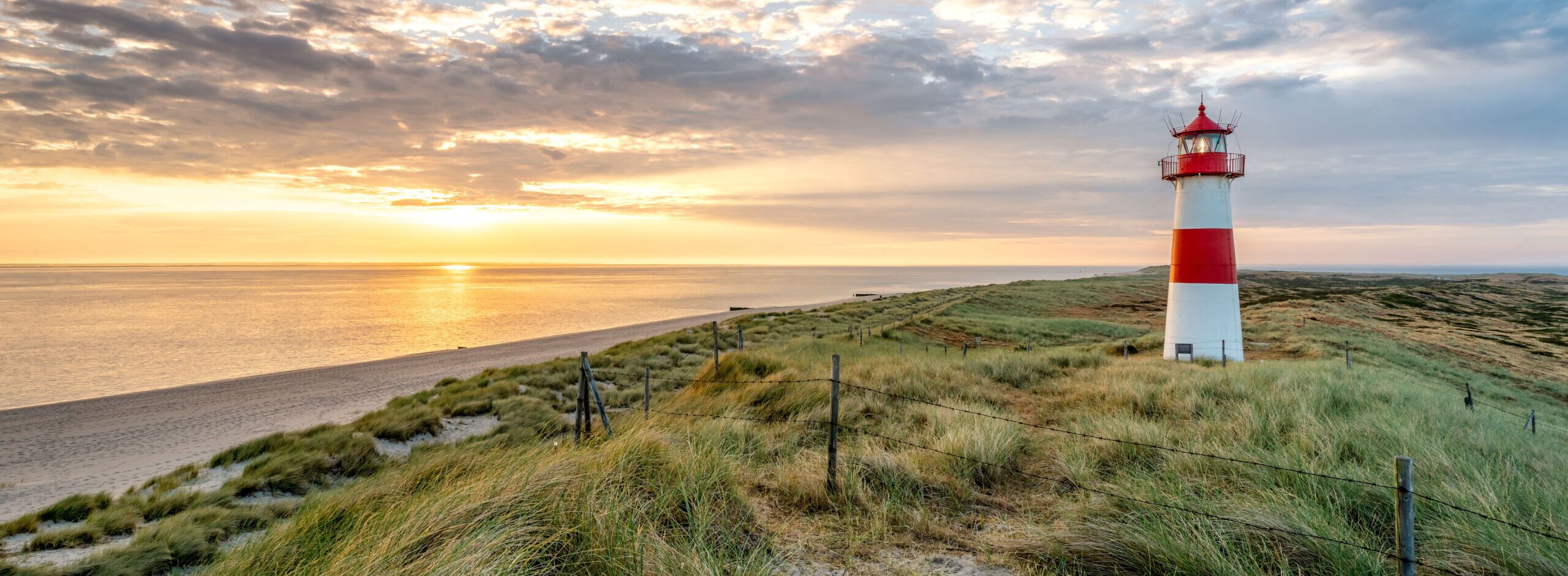 Sonnenuntergang am Strand in Sylt. Im Vordergrund Dünen und ein rot-weißer Leuchtturm