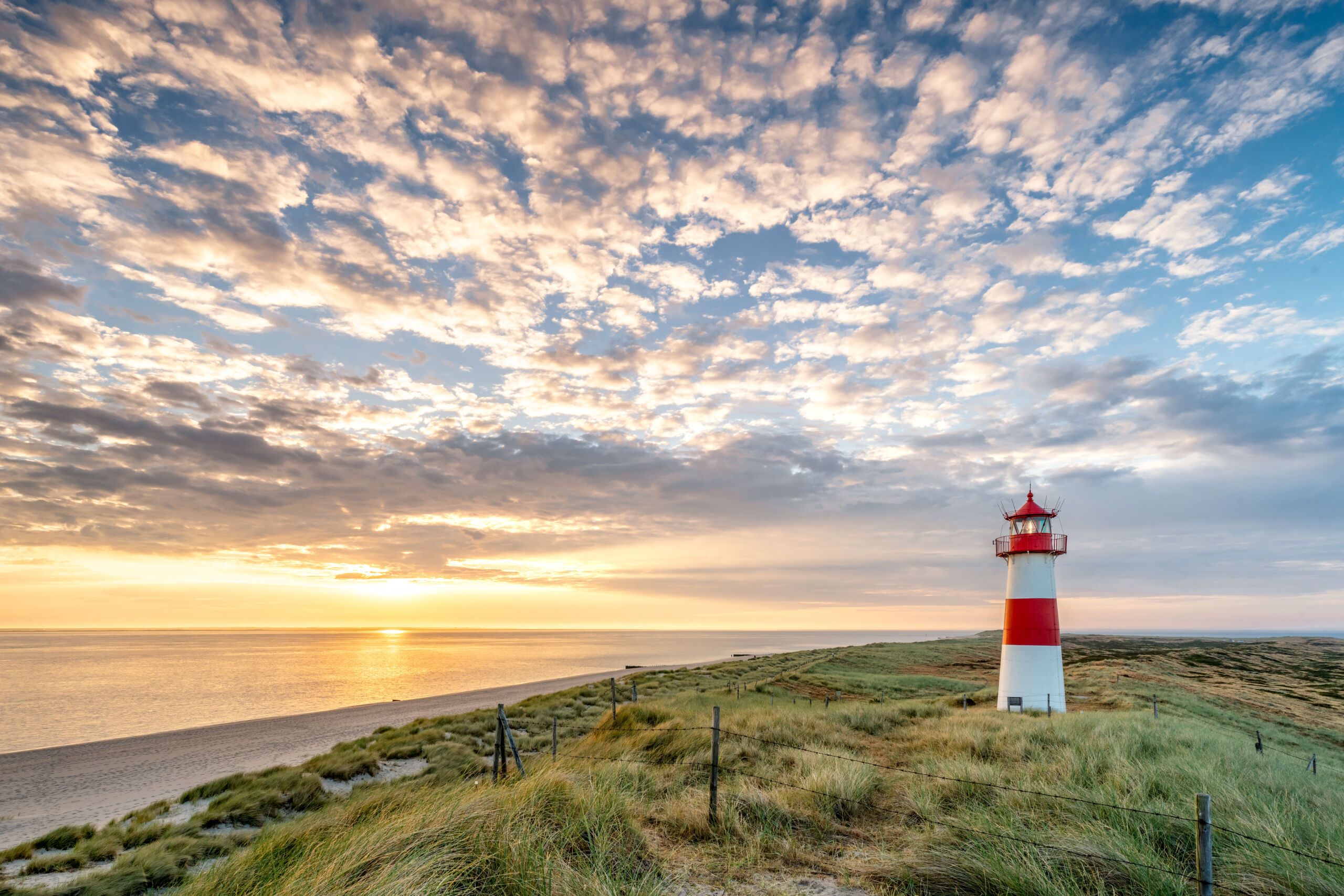 Sonnenuntergang am Strand in Sylt. Im Vordergrund Dünen und ein rot-weißer Leuchtturm