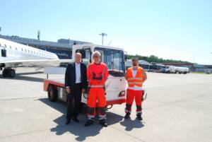 Elektroschlepper auf dem Vorfeld des Flughafens. Davor stehen drei männliche Personen. Zwei davon in Arbeitskleidung und einer im Anzug
