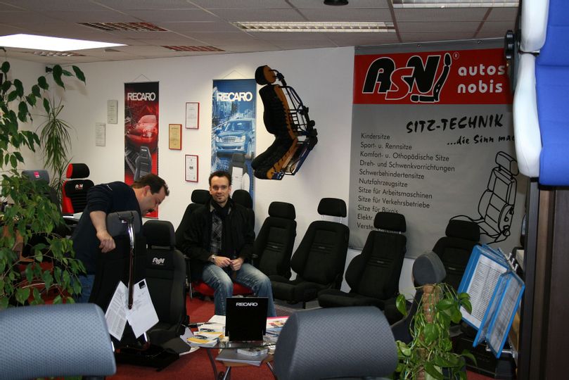 Innenbereich von ASN Autositze in Bad Wildungen. Zu sehen sind mehrere schwarze Autositze. Auf einem davon sitzt ein Mann. Links daneben schaut sich ein anderer Mann einen Sitz von hinten genauer an.
