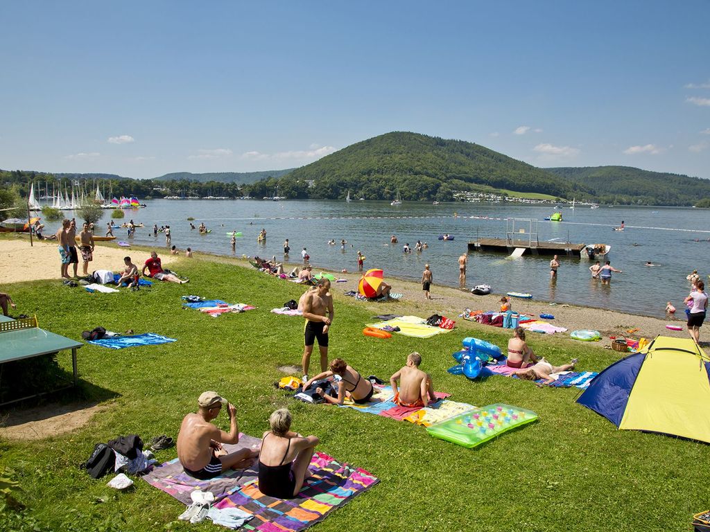 Personen haben sich am Ufer des Edersees mit Handtüchern ausgebreitet. Viele sind am schwimmen.