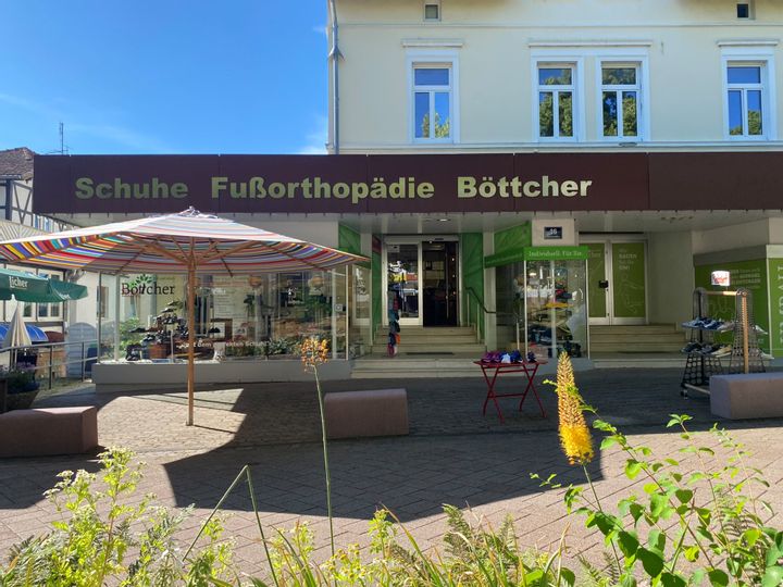 Außenansicht des Eingangs von Schuhe und Fußorthopädie Böttcher. Vor dem Eingang ist eine Treppe. Es stehen ein bunter Sonnenschirm sowie ein Regal und ein Tisch vor dem Eingang mit Schuhen.