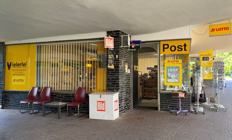 Außenansicht des Eingangs von Heinzers Geschenkpost. Vor dem Eingang links sind drei Stühle und ein Tisch zu sehen und auf der rechten Seite sind Ständer mit Ansichtskarten.
