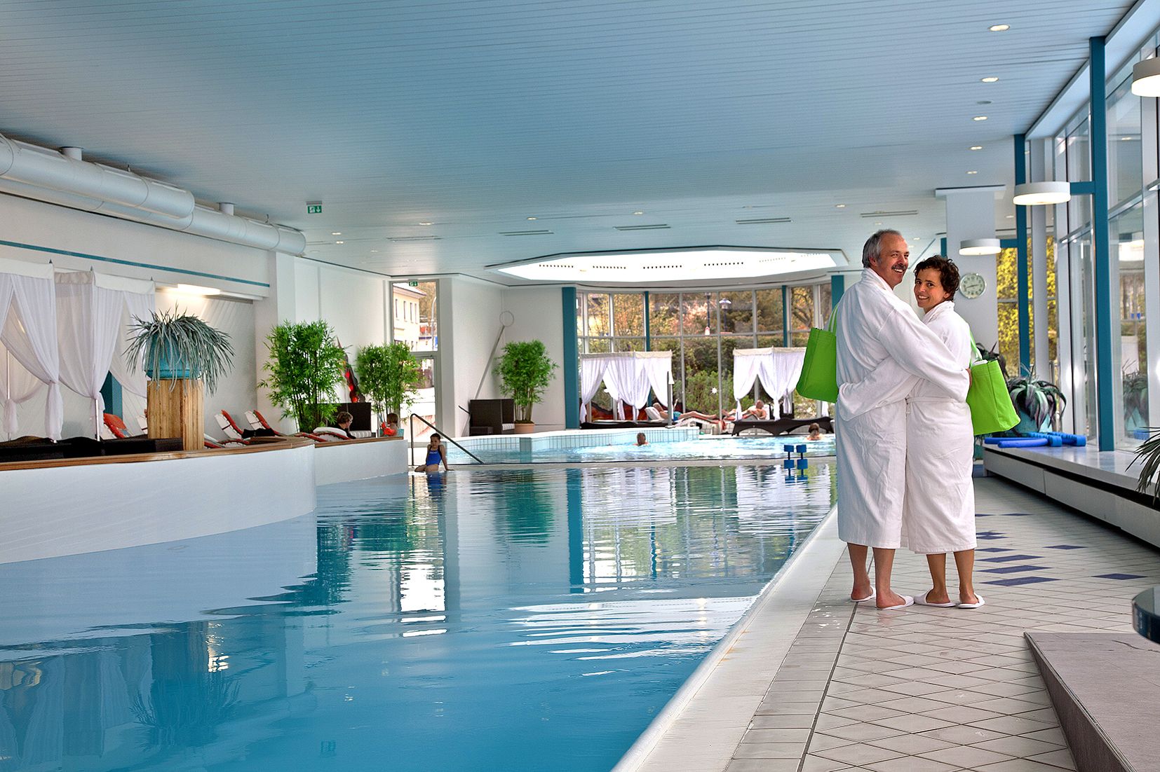 Ein Mann und eine Frau, die sich im Bademantel im Arm halten undneben dem Schwimmbecken der Quellentherme Reinhardshausen stehen und in die Kamera schauen.