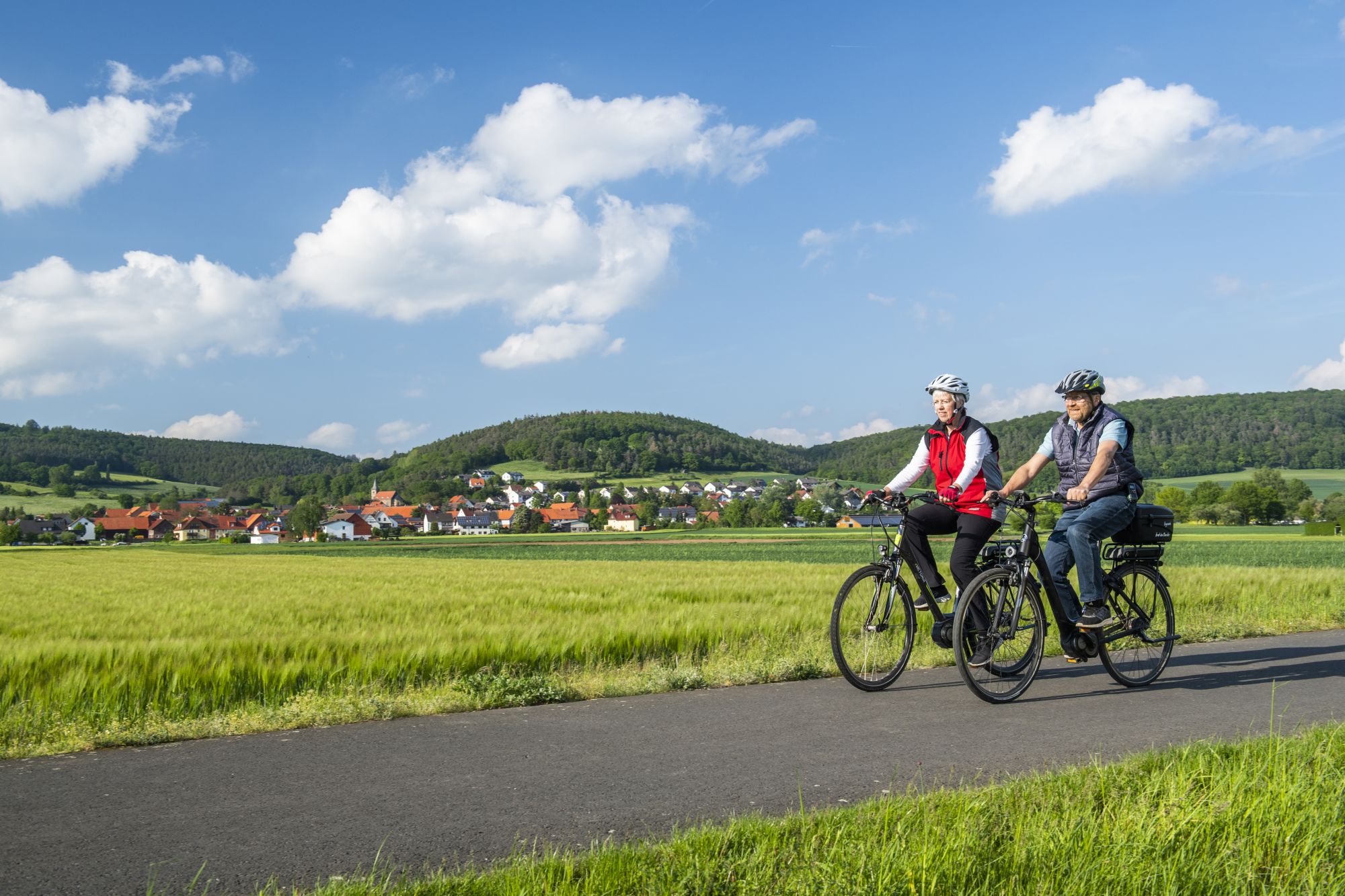 Zwei Radfahrer fahren auf einen Feldweg neben grünen Feldern und einem Dorf im Hintergrund