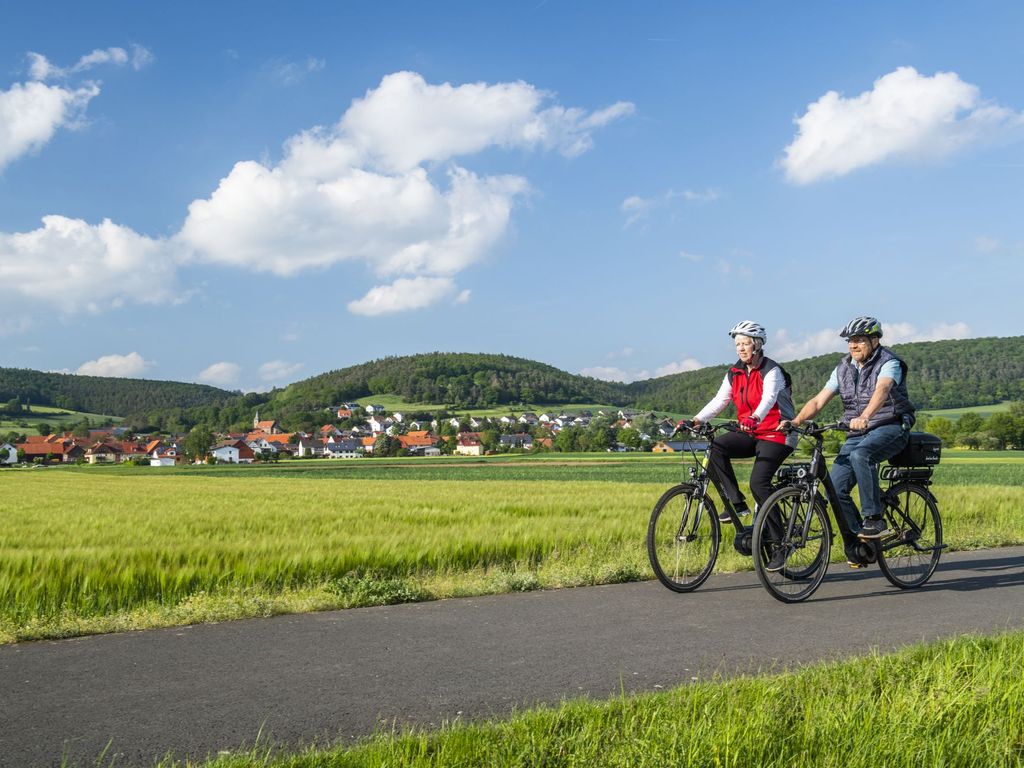 Zwei Radfahrer fahren auf einen Feldweg neben grünen Feldern und einem Dorf im Hintergrund