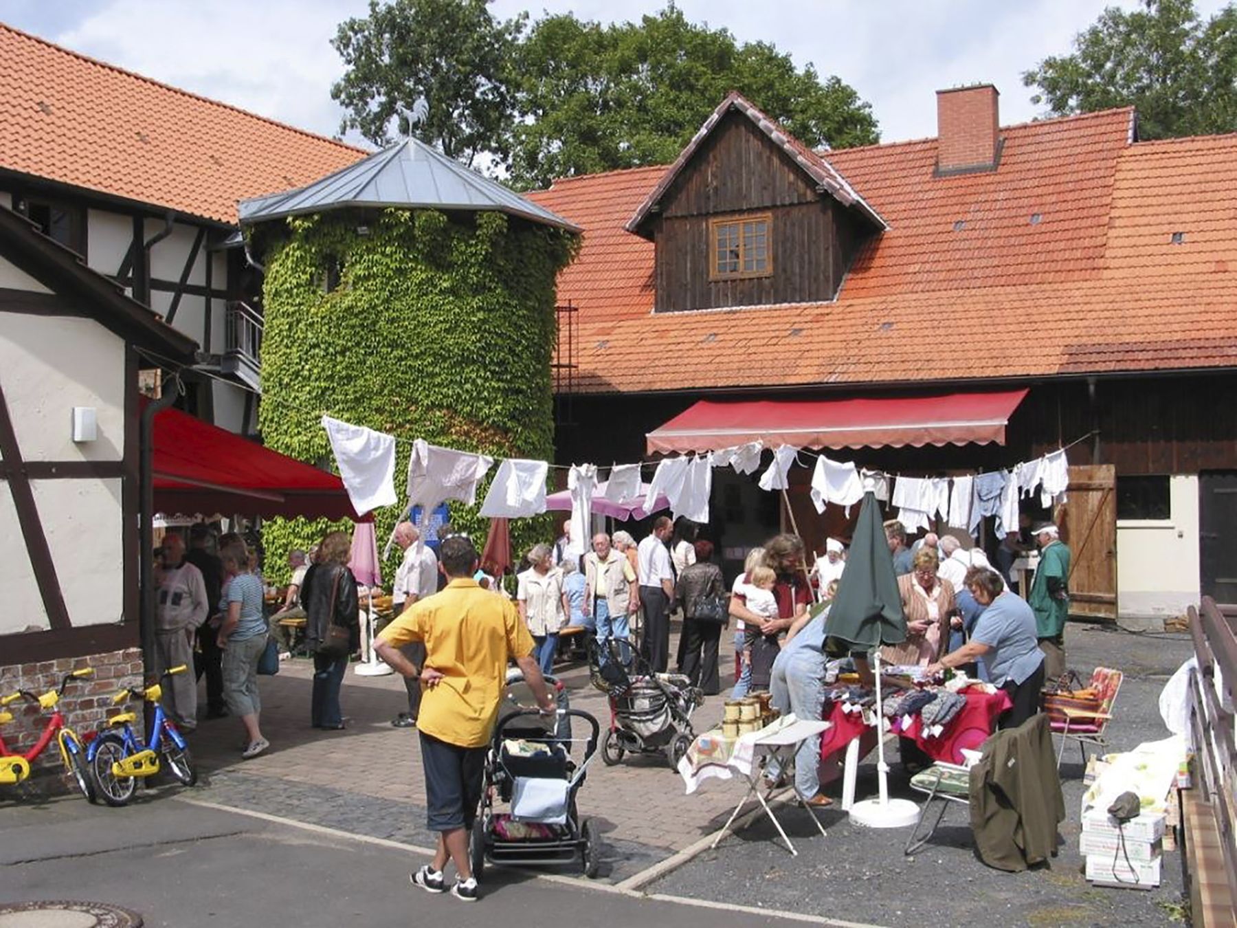Blick auf den Hof des Lebendigen Museums in Oderhausen mit einer Gruppe von Menschen und einem Marktstand,