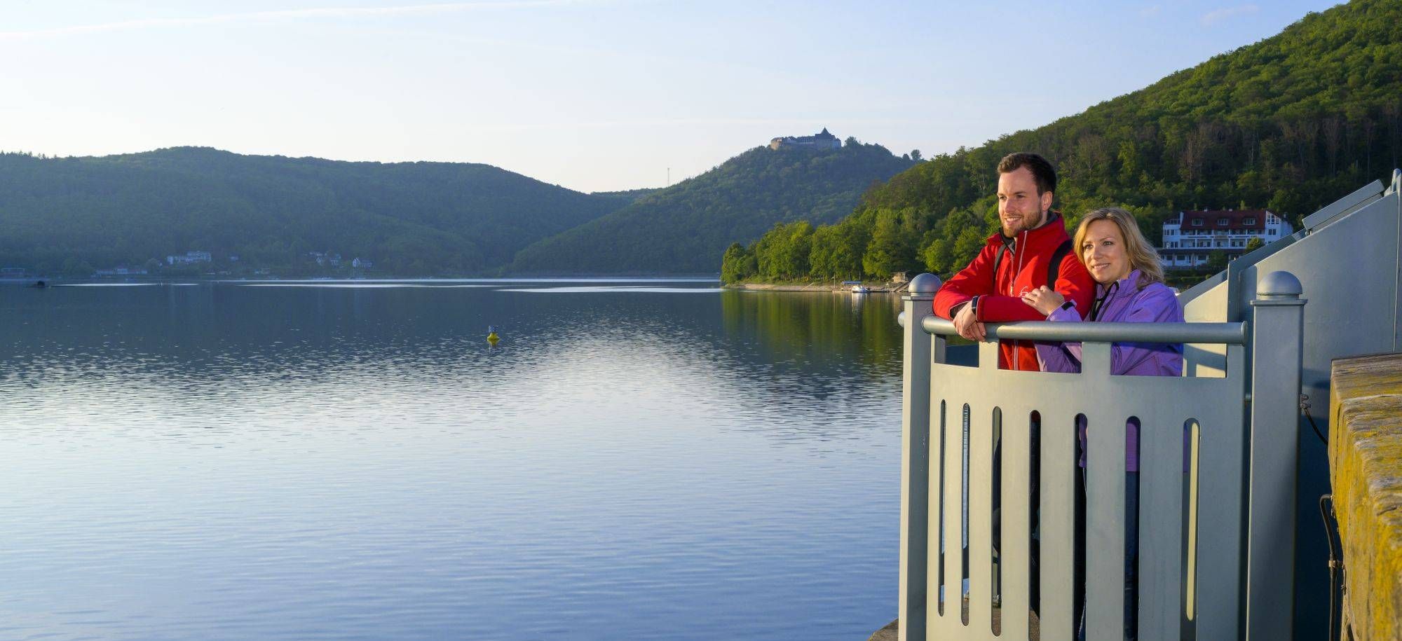 Zwei Personen am Ausguck schauen auf den Edersee. Im Hintergrund ist der Edersee mit Hügeln und Bergen.