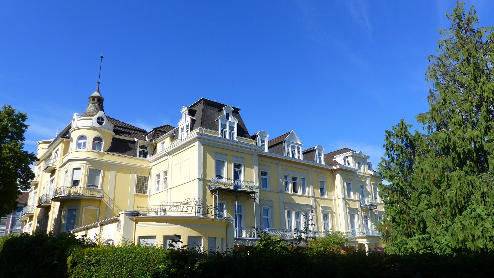 Aufnahme des Kaiserhofs in Bad Wildungen. Im Vordergrund ist eine grüne Hecke. Rechts ist ein Nadelbaum. Der Himmel ist blau mit einzelnen Wolken.