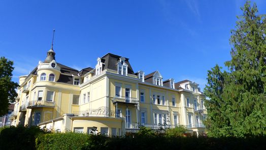 Aufnahme des Kaiserhofs in Bad Wildungen. Im Vordergrund ist eine grüne Hecke. Rechts ist ein Nadelbaum. Der Himmel ist blau mit einzelnen Wolken.