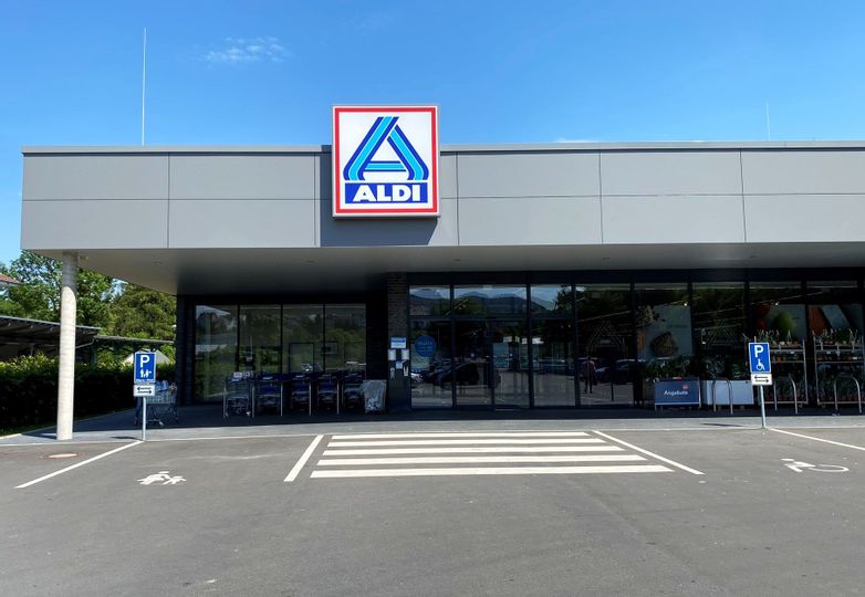 Außenansicht des Eingangs von Aldi. Vor dem Gebäude sind Parkplatze und neben dem Eingang die Einkaufswagen zu sehen.