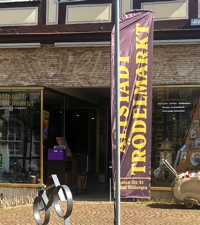 Außenansicht des Eingangs vom Altstadt Trödelmarkt. Vor dem Eingang steht eine Fahne mit dem Namen des Geschäfts und rechts davon ein alter Puppenwagen.