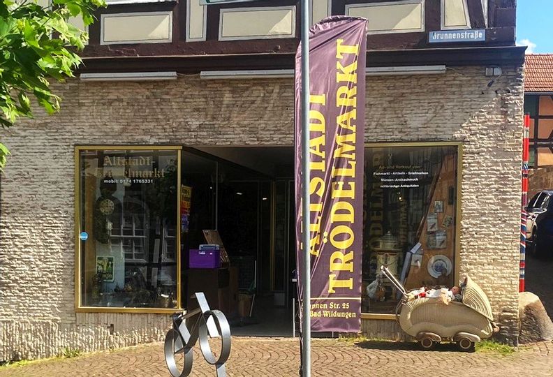 Außenansicht des Eingangs vom Altstadt Trödelmarkt. Vor dem Eingang steht eine Fahne mit dem Namen des Geschäfts und rechts davon ein alter Puppenwagen.