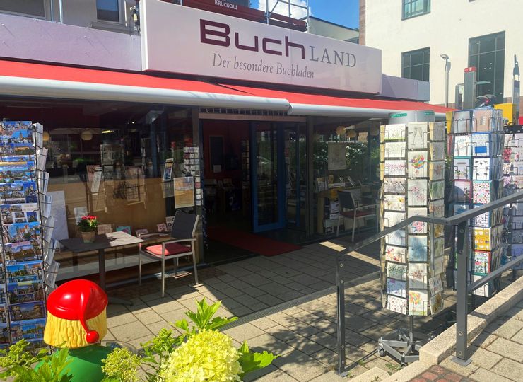 Außenansicht des Eingangs von Buchland der besondere Buchladen. Rechts und links im Bild sind Ständer mit Ansichtskarten zu sehen und neben dem Eingang Tische und Stühle.
