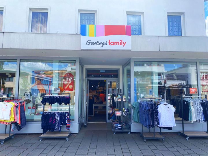 Außenansicht des Eingangs von Ernsting´s family. Rechts und links von dem Eingang stehen Ständer mit Kinder und Damenbekleidung.