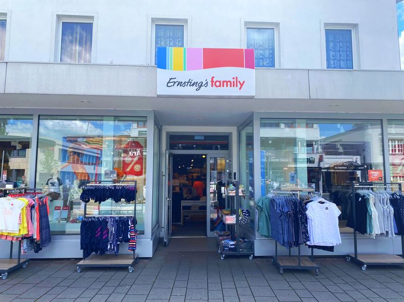 Außenansicht des Eingangs von Ernsting´s family. Rechts und links von dem Eingang stehen Ständer mit Kinder und Damenbekleidung.