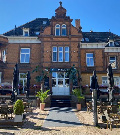 Außenansicht der Restaurant-Bar Havana mit dem Eingangsbereich und er Terrasse vor dem Gebäude bei sonnigem Wetter.