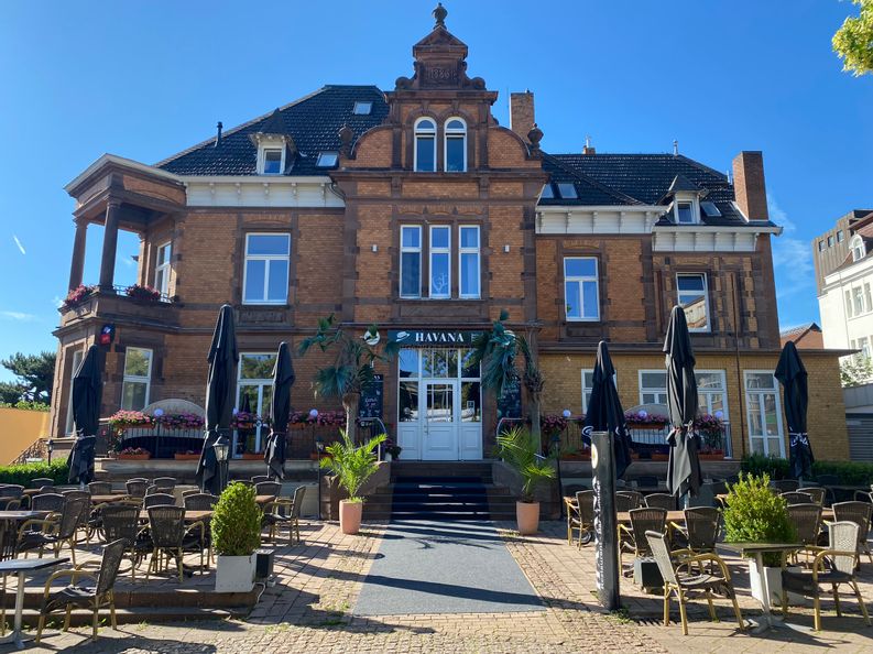 Außenansicht der Restaurant-Bar Havana mit dem Eingangsbereich und er Terrasse vor dem Gebäude bei sonnigem Wetter.
