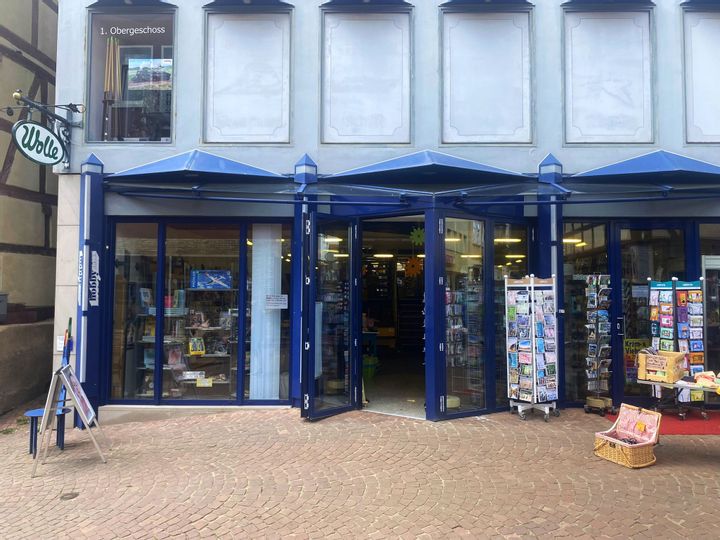 Außenansicht des Eingangs vom Hobbyhaus Hommel. Die Tür und Fensterrahmen sind blau und rechts vom Eingang sind Ständer mit Geschenkarten zu sehen.