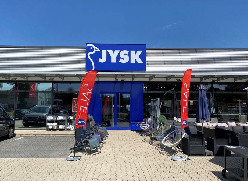 Außenansicht des Eingangs von Jysk. Links vom Eingang sind Parkplätze und rechts vom Eingang sind Gartenstühle zu sehen.