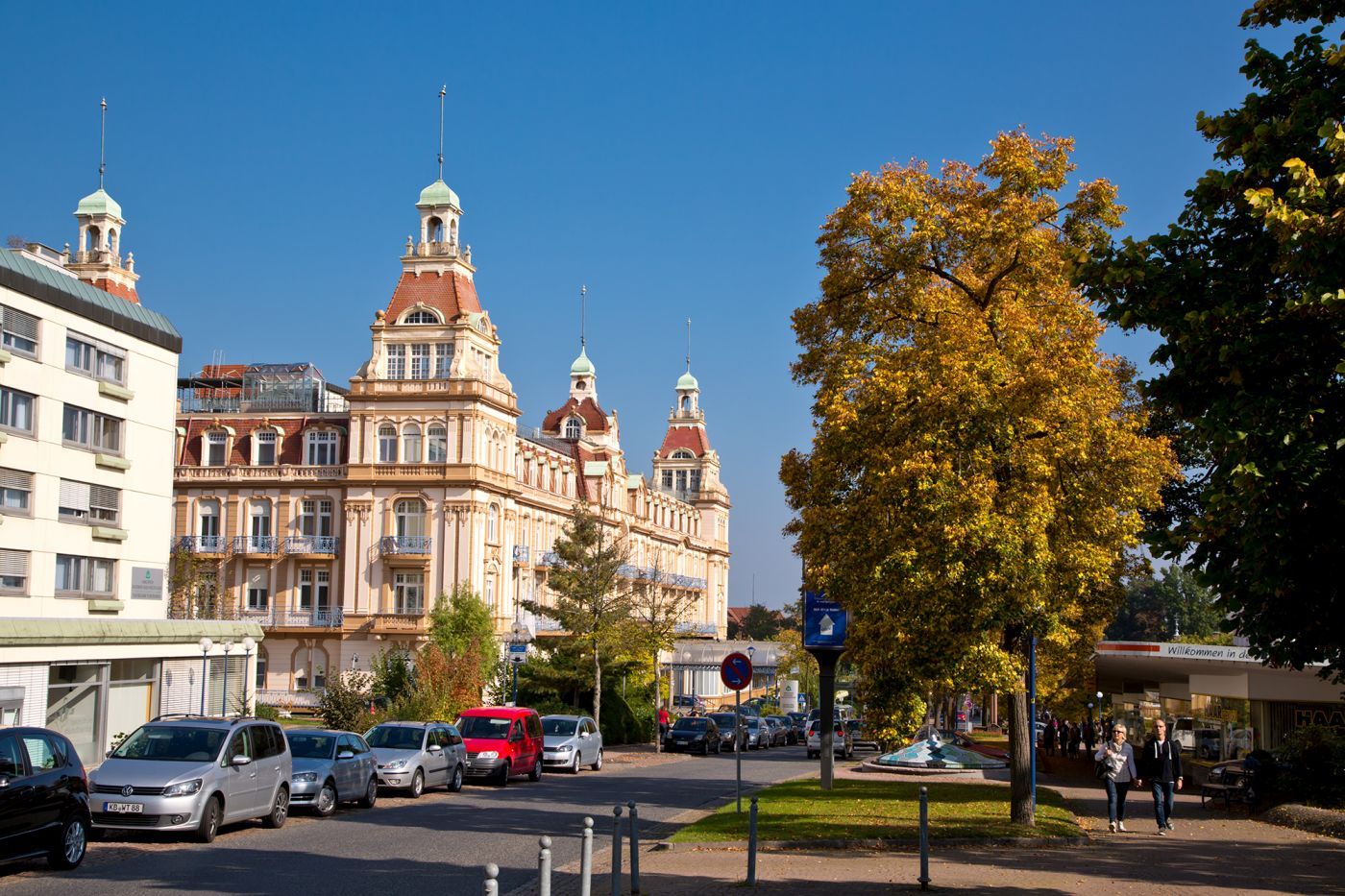 Blick auf die Brunnenallee und den Fürstenhof im Herbst bei sonnigem Wetter. Auf der Brunnenallee stehen viele Autos am Straßenrand und rechts im Bild sind Spaziergänger zu sehen.