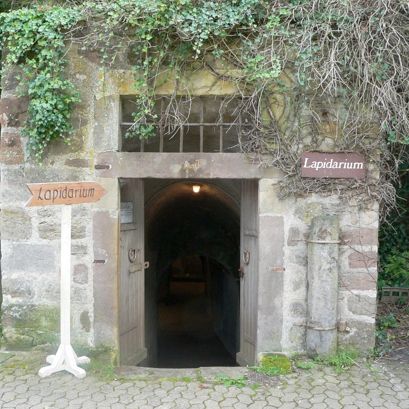 Außenansicht des Eingangs des Lapidarium am Schloss Friedrichstein in Bad Wildungen. Recht und links von dem Eingang sind jeweils ein Schild mit dem Wort Lapidarium zu sehen. Über dem Eingang hängen Sträucher herunter.