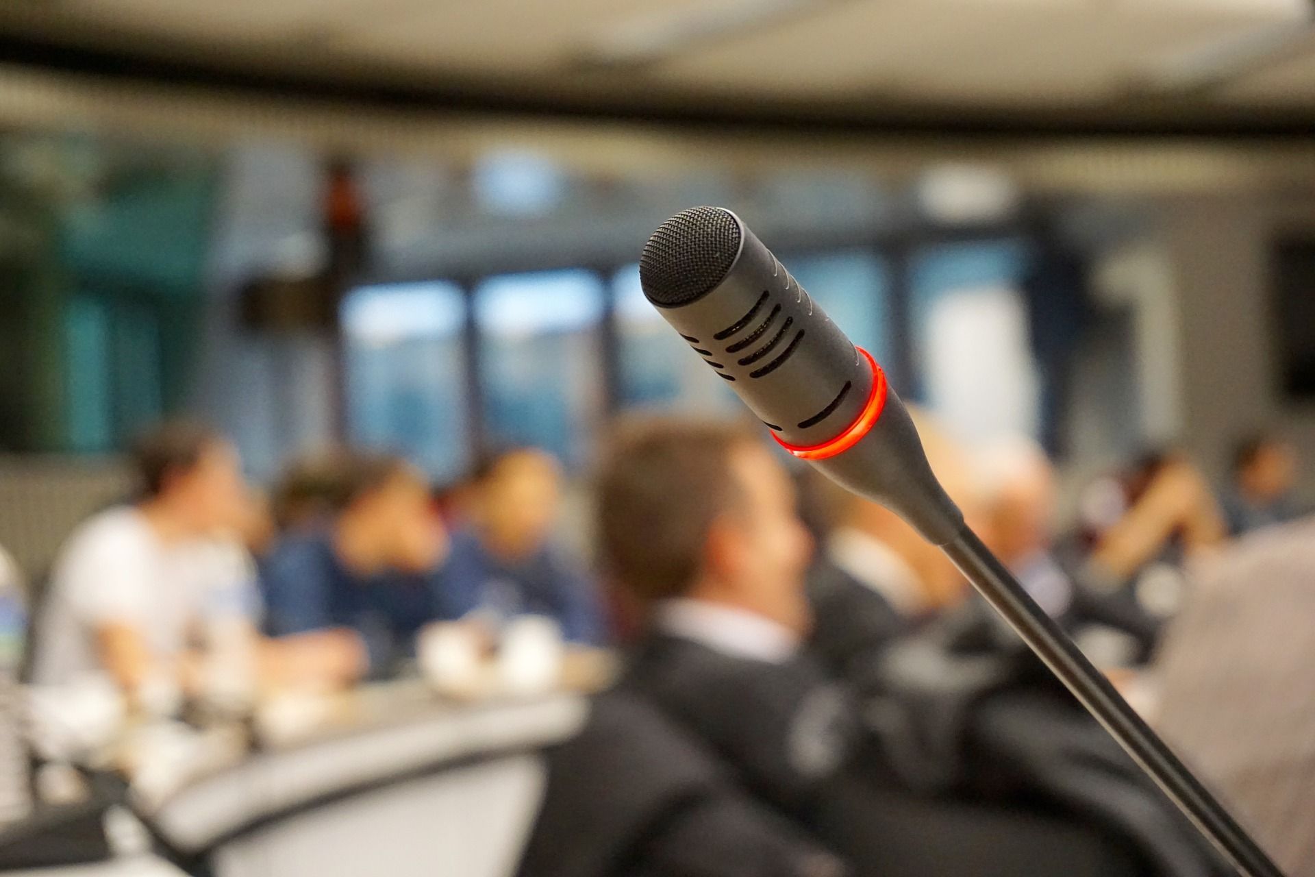 Im Vordergrund befindet sich ein schwarzes Mikrofon. Im Hintergrund sitzen Personen in einem Saal.