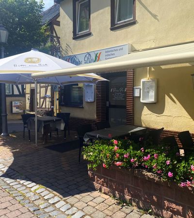 Eingang des Pizza Pie in Bad Wildungen bei sonnigem Wetter. Es ist ein Sonnenschirm sowie eine Markise zu sehen unter denen Tische und Stühle stehen.