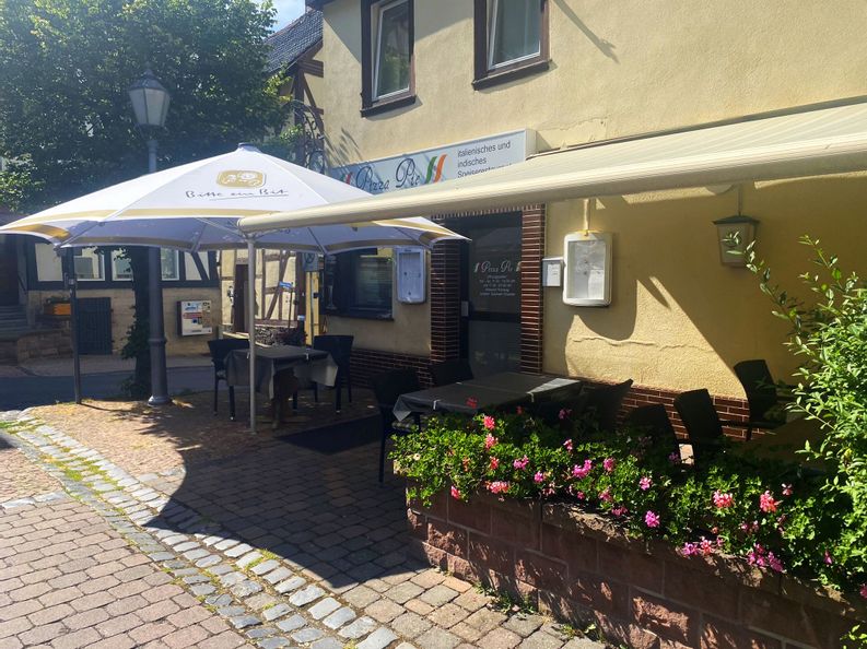 Eingang des Pizza Pie in Bad Wildungen bei sonnigem Wetter. Es ist ein Sonnenschirm sowie eine Markise zu sehen unter denen Tische und Stühle stehen.