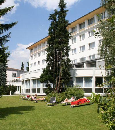 Außenansicht der Klinik Reinhardsquelle. Vor dem Gebäude ist eine Wiese mit Bäumen zu sehen. Sechs liegen stehen auf der Wiese auf denen Menschen liegen.