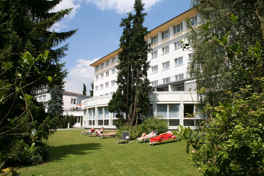 Außenansicht der Klinik Reinhardsquelle. Vor dem Gebäude ist eine Wiese mit Bäumen zu sehen. Sechs liegen stehen auf der Wiese auf denen Menschen liegen.