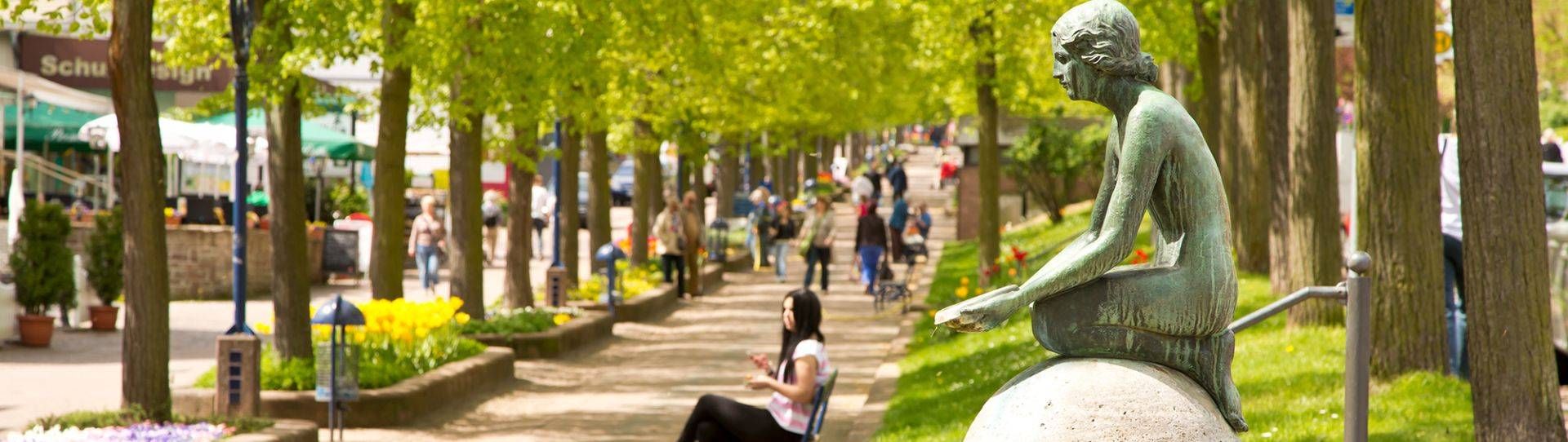 Blick entlang der Brunnenallee in Bad Wildungen mit dem Nymphenbrunnen im Vordergrund und laufenden Menschen im Hintergrund.