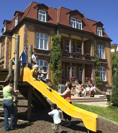 Blick auf das Gebäude der Mutter-Kind-Klinik Talitha. Vor dem Gebäude ist ein Spielplatz zu sehen. Einige Kinder rutschen auf einer gelben Rutsche. Zwei Mütter stehen daneben. Im Hintergrund sieht man weitere Mütter auf einer Bank sitzen.