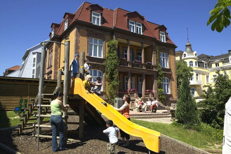 Blick auf das Gebäude der Mutter-Kind-Klinik Talitha. Vor dem Gebäude ist ein Spielplatz zu sehen. Einige Kinder rutschen auf einer gelben Rutsche. Zwei Mütter stehen daneben. Im Hintergrund sieht man weitere Mütter auf einer Bank sitzen.