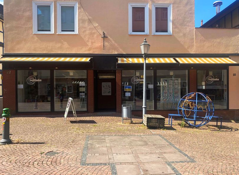 Außenansicht des Eingangs vom Weltladen. Vor dem Eingang steht eine blau-gelbe Weltkugel aus Metall sowie ein Bank aus Holz.