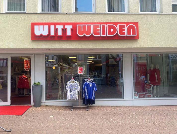 Außenansicht des Eingangs des Geschäfts Witt Weiden. Vor dem Eingang steht ein Ständer mit T-Shirts.