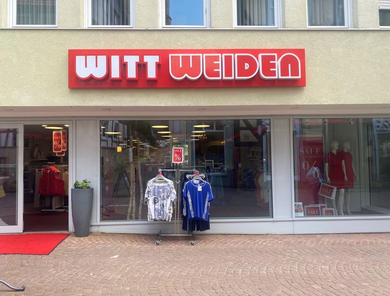 Außenansicht des Eingangs des Geschäfts Witt Weiden. Vor dem Eingang steht ein Ständer mit T-Shirts.