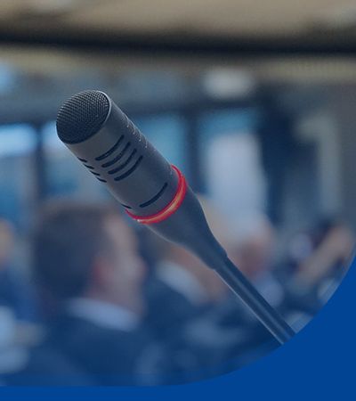 Blick auf ein schwarzes Mikrofon. Im Hintergrund sitzen Personen in einem Saal