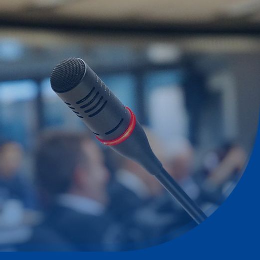 Blick auf ein schwarzes Mikrofon. Im Hintergrund sitzen Personen in einem Saal