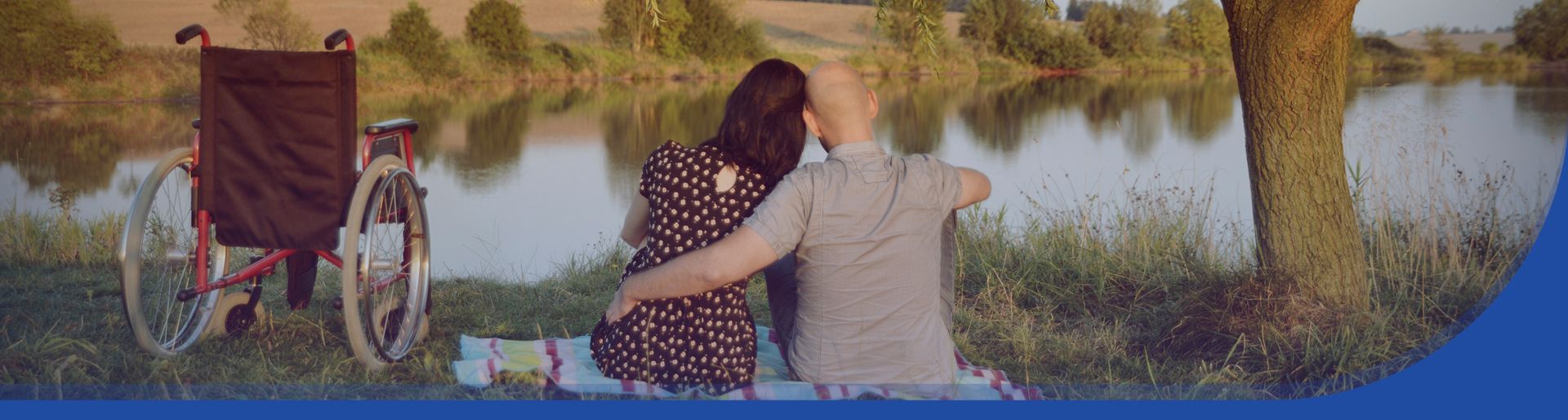 2 Personen sitzen zusammen am See und blicken in die Ferne. Links von ihnen ein Rollstuhl