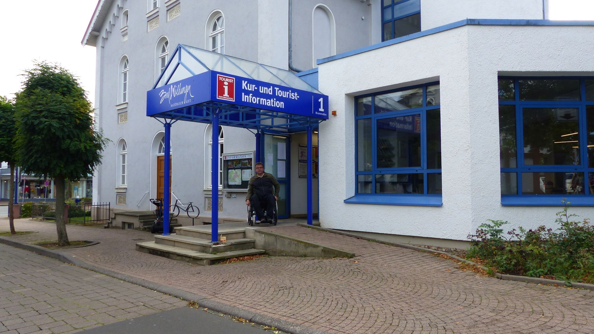 Barrierefreiheit am Beispiel der Tourist-Information in Bad Wildungen. Ein Rollstuhlfahrer ist am Eingang der Tourist-Information und kann über die barrierefreie Rampe ins innere des Gebäudes gelangen.