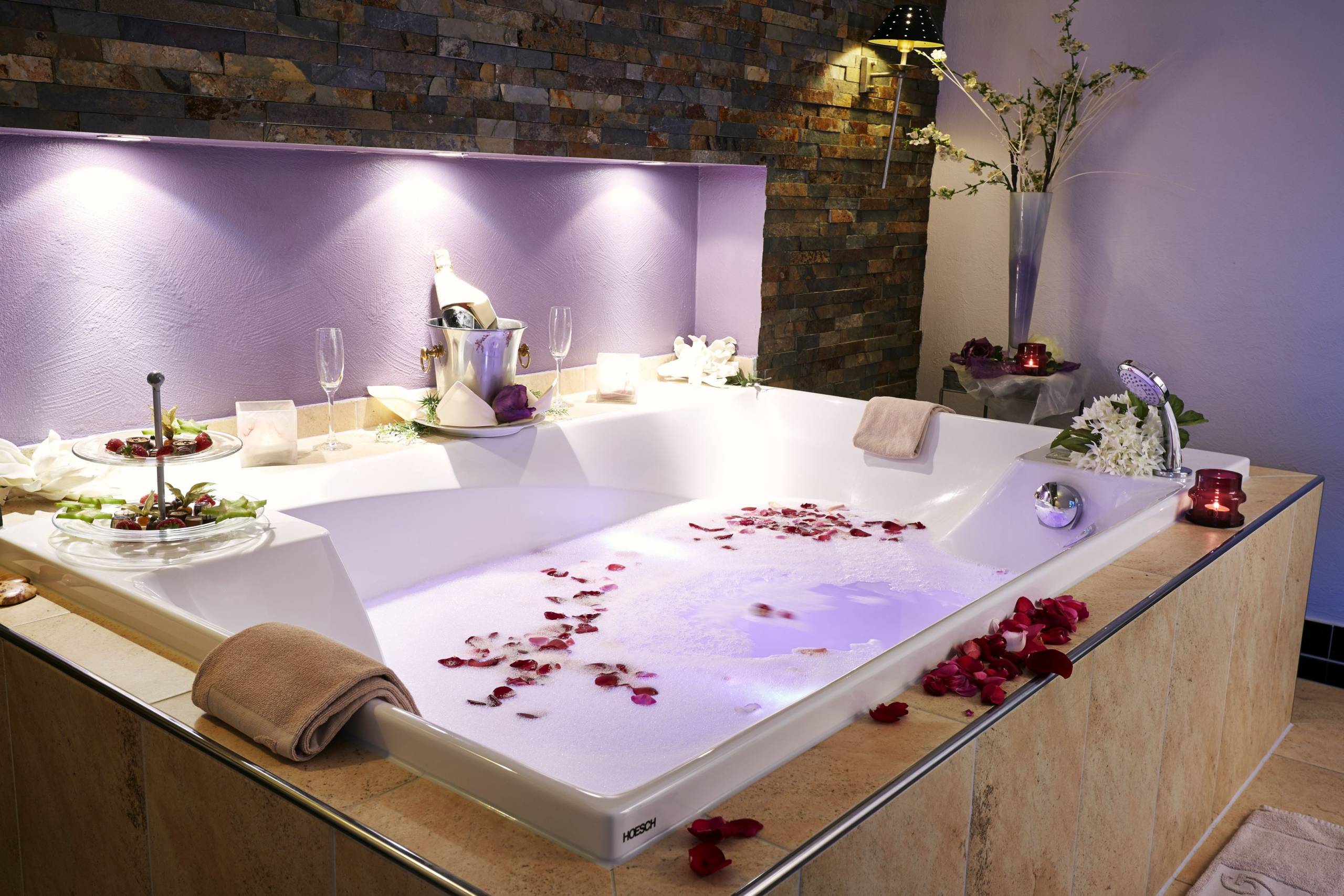 Wellnessbereich und Badewanne im Goebel's Hotel AquaVita. Gefüllt mit Wasser und Rosenblättern. Auf dem Rand stehen weiße Blumen, Sektgläser, Sekt und ein Teller mit Snacks.