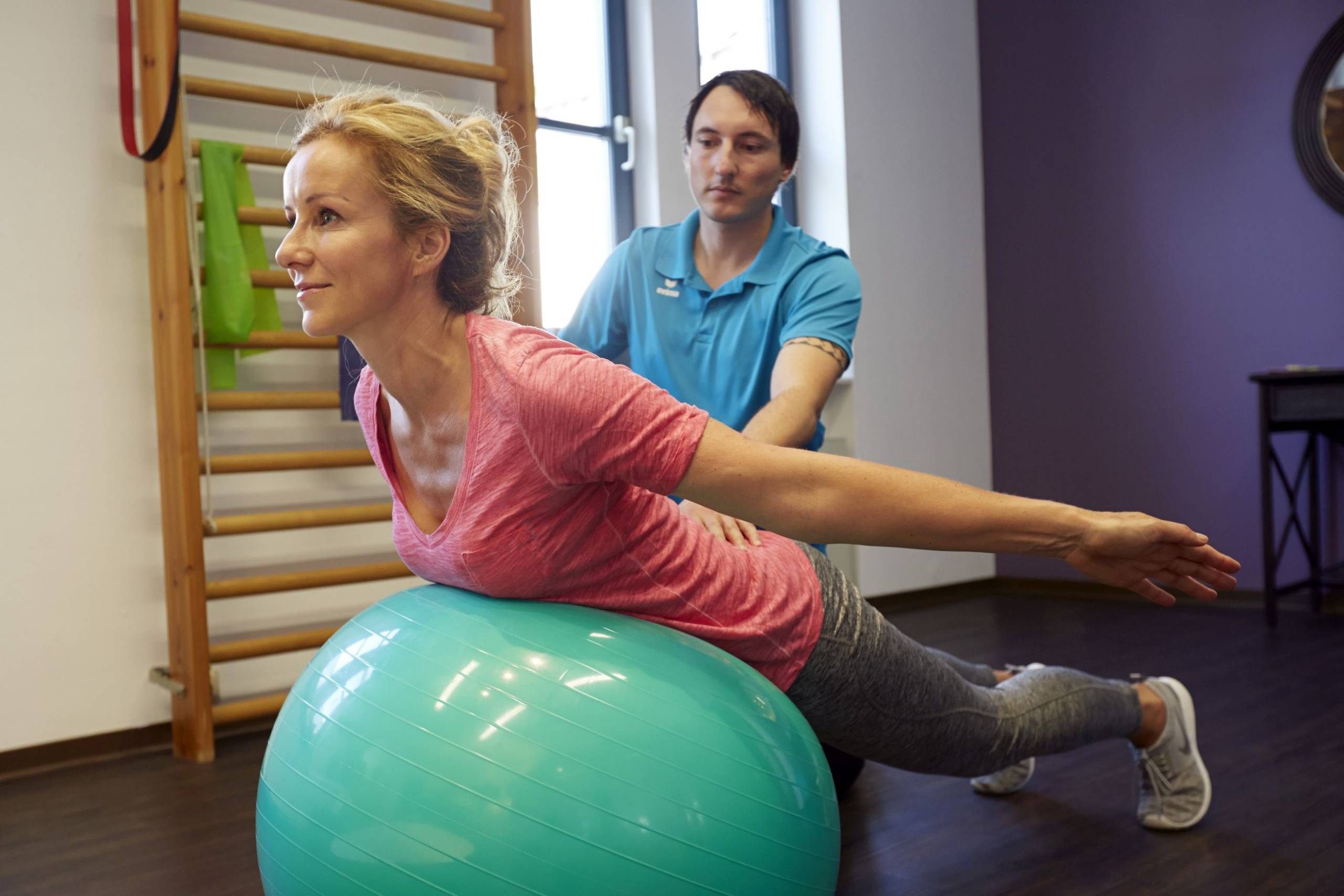 Eine Frau lieg mit dem Bauch auf einem grünen Gymnastikball. Hinter ihr hockt ein Physiotherapeut, der ihr bei den Übungen hilft.