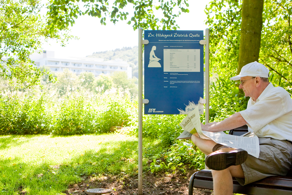 Dr. Hildegard Dietrich Quelle in Bad Wildungen in freier Natur bei sonnigem Wetter. Ein Mann sitzt auf einer Bank neben dem Quellenschild und liest eine Zeitung.