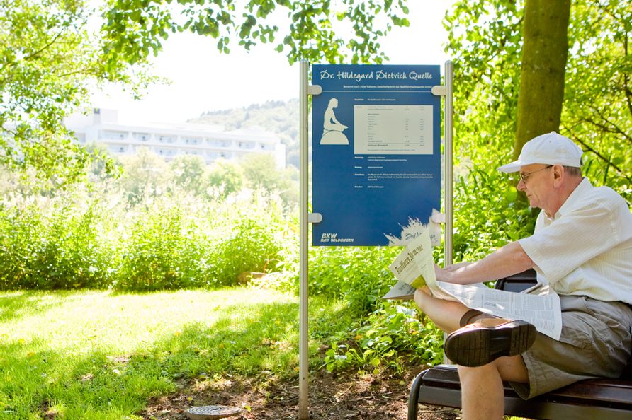 Dr. Hildegard Dietrich Quelle in Bad Wildungen in freier Natur bei sonnigem Wetter. Ein Mann sitzt auf einer Bank neben dem Quellenschild und liest eine Zeitung.