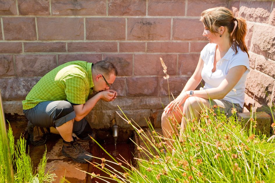 Ein Mann trinkt aus einer Quelle im Freien und eine Frau sitzt rechts daneben und schaut zu.