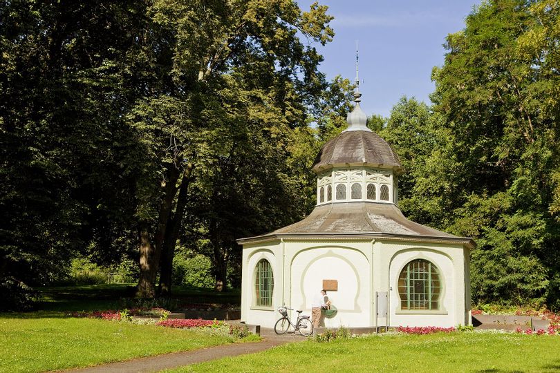 Blick auf das Gebäude der Königsquelle im Bad Wildunger Kurpark umgeben von Bäumen bei sonnigem Wetter. Vor dem Häuschen ist ein Fahrrad vor den Außenzapfstelle abgestellt.
