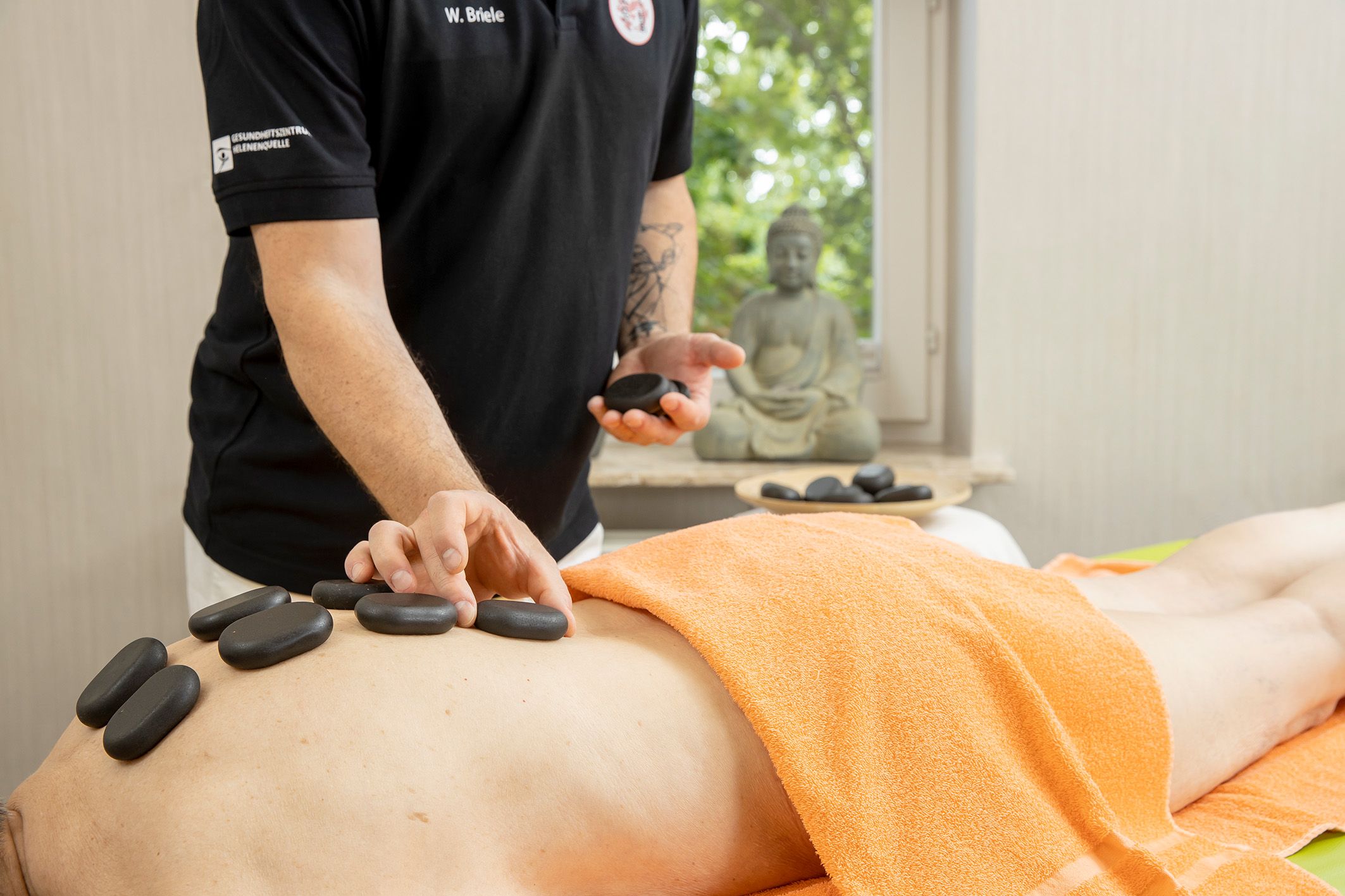 Gesundheitszentrum Helenenquelle. Eine Person liegt auf einer Liege. Auf den Rücken werden von einem Therapeuten schwarze Steine gelegt.