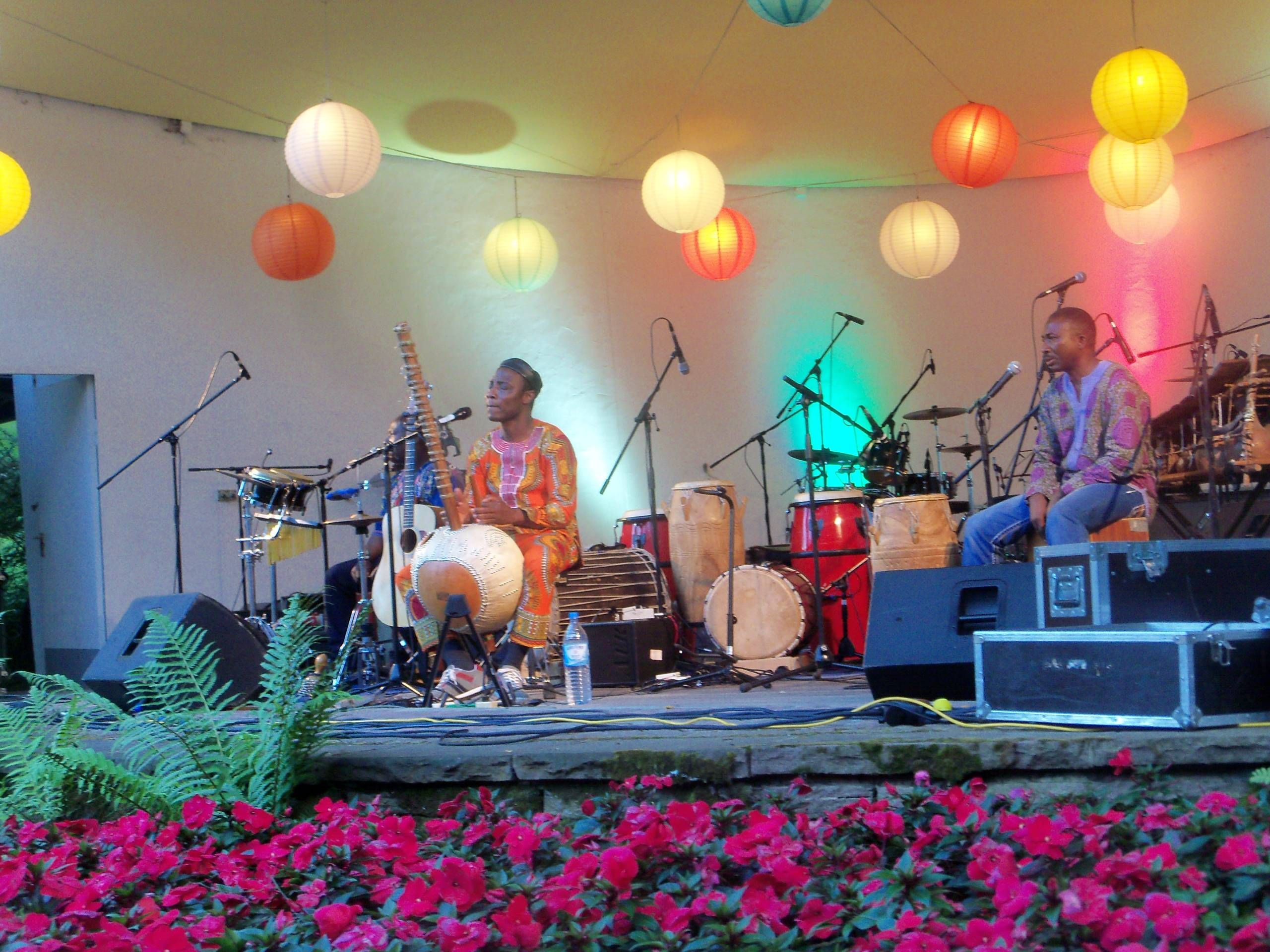 Eine Band spielt im Rahmen des Folk im Park im Musikpavillon der Wandelhalle Bad Wildungen. An der Decke des Pavillons hängen Lampions. Auf der Bühne sitzen zwei Künstler mit vielen Instrumenten und vor dem Musikpavillon blühen viele pinke Blumen.